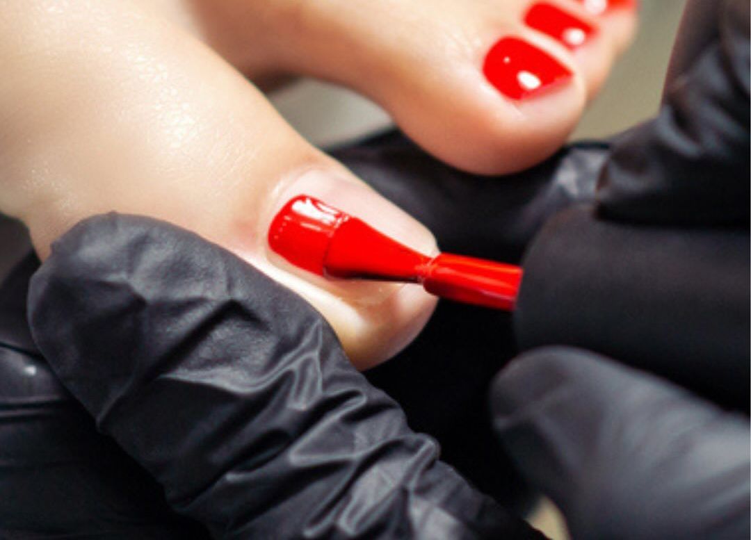 Nail technician in black gloves painting a pedicure red