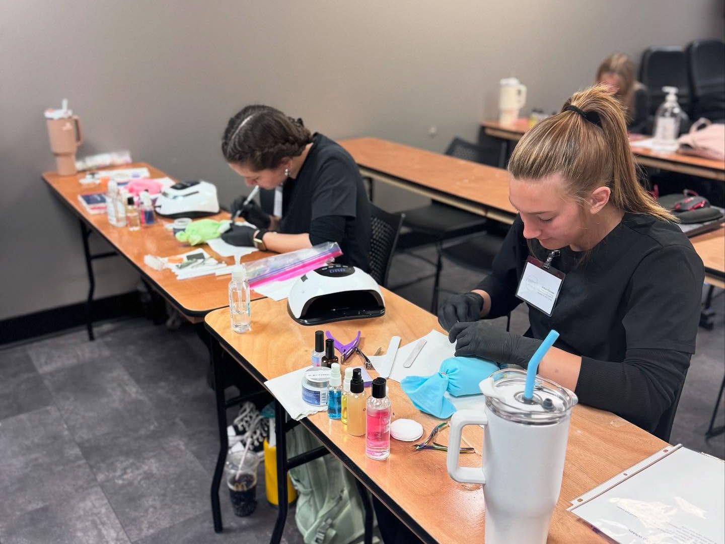 Two students in black scrubs and gloves practicing manicures at classroom tables